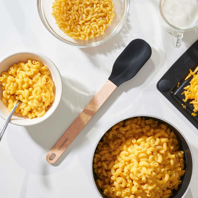 Epicurean Silicone Series Small Spoonula in Natural/Black on a white counter surrounded with bowls of ready to eat macaroni and cheese with all of the ingredients to prepare more