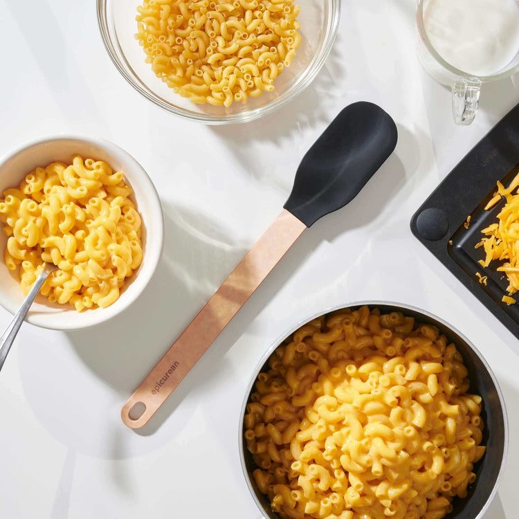 Epicurean Silicone Series Small Spoonula in Natural/Black on a white counter surrounded with bowls of ready to eat macaroni and cheese with all of the ingredients to prepare more