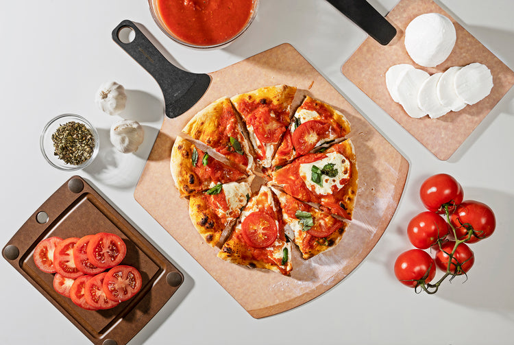 Pizza Peel Plus featured with a cheese and tomato pizza, along side the Handy Series Plus in Natural rectangle with cheese and an All-In-One Cutting Board in Natural with tomatoes 